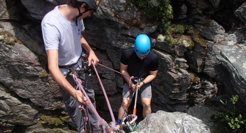 Two people wearing safety gear are secured by ropes as they prepare to rappel down a cliff. One person appears to be an instructor, giving direction to the other person.